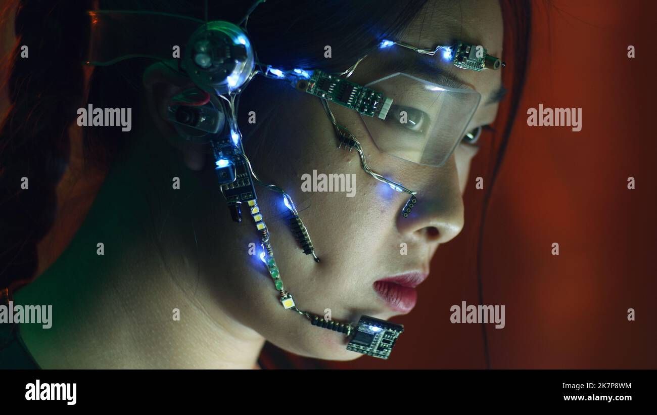 Fokussiertes asiatisches Mädchen in Cyberpunk-Kleidung arbeitet am Computer. Tragen einer einäugigen Brille mit weißen LED-Leuchten und einem Mikrofon. Neonlicht Hintergrund. Science Fiction, Kybernetik-Konzept. Stockfoto