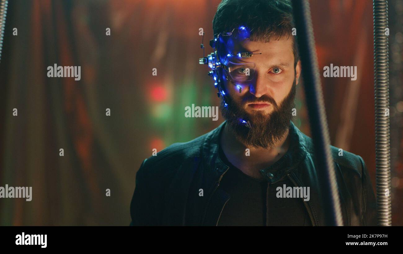 Der junge Mann mit einem Headset mit LED-Leuchten steht hinter den schwarzen Hängestangen und schaut auf die Kamera. Cyberpunk und futuristisches Konzept. Neonlichter im Hintergrund. Stockfoto