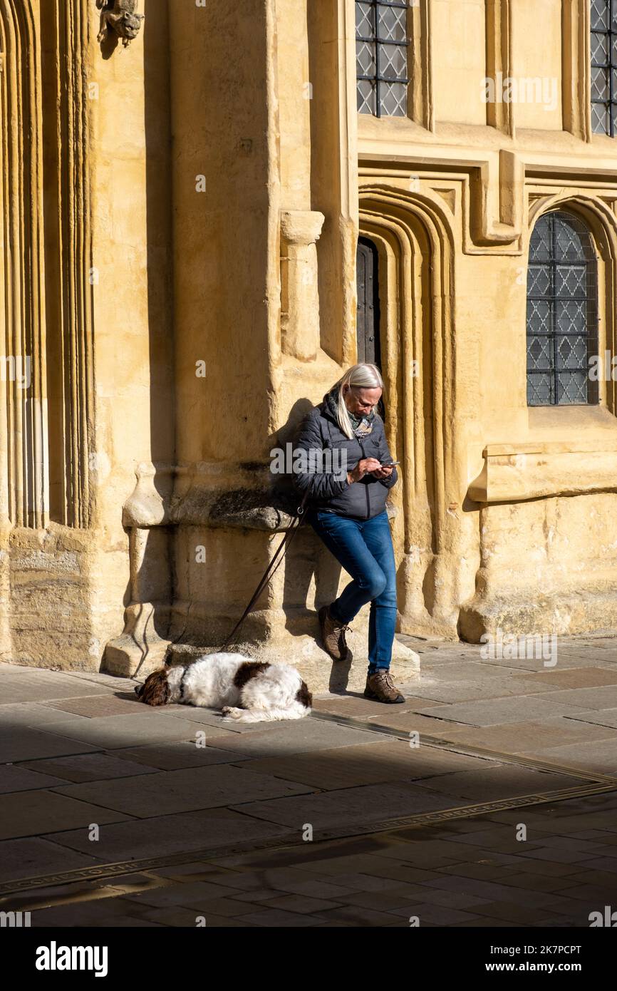 Reife Frau, die ihr Telefon gegen den Eingang zur St. gelehnt Johannes-Täufer-Kirche in Cirencester (Oct22) Stockfoto