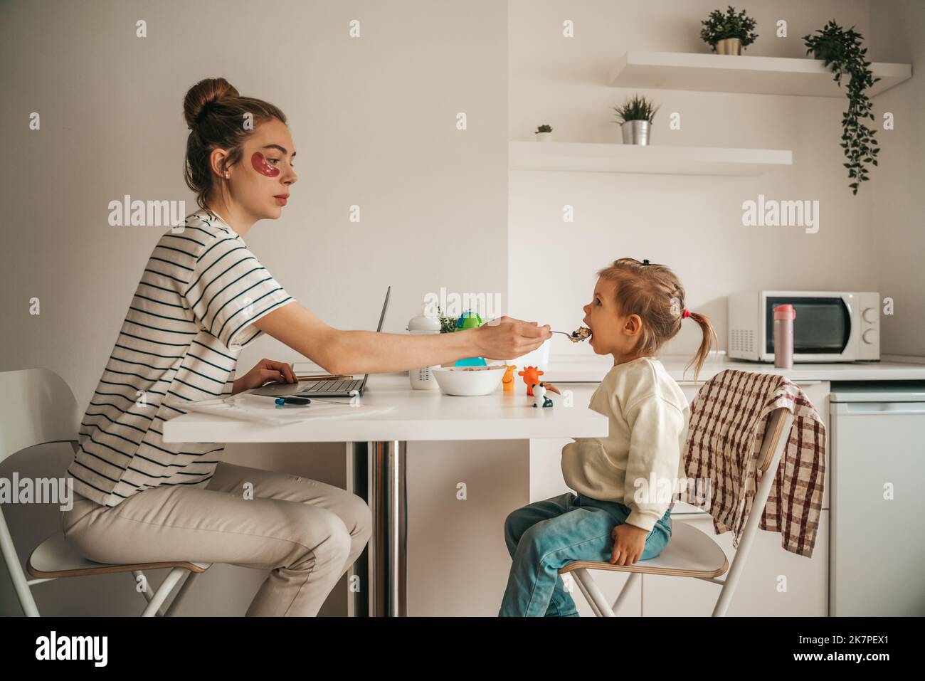 Frau mit Unteraugenflecken sitzt vor dem Laptop und gibt dem Mädchen am Küchentisch einen Löffel Haferbrei Stockfoto