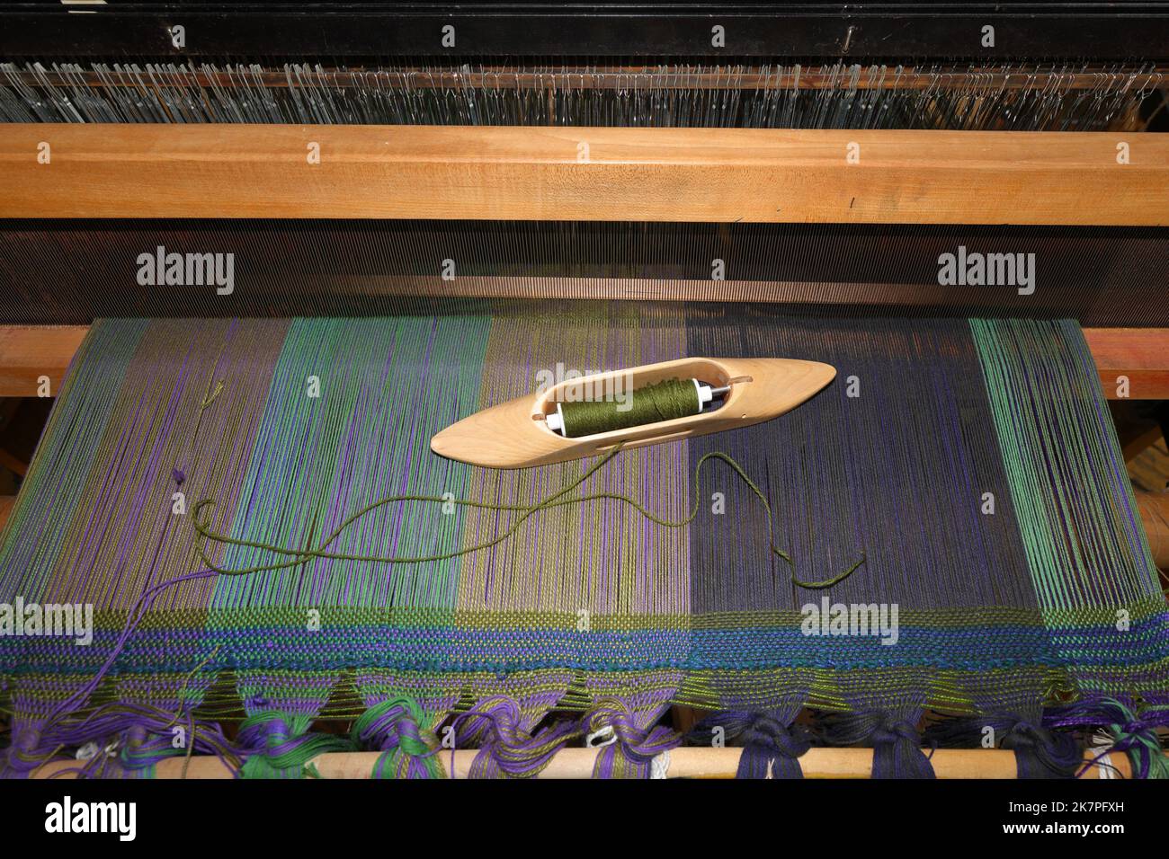 Ein Bootsshuttle mit offenem Boden und einer Spule aus grünem Garn, die auf einem Webstoff auf einem Webstuhl ruht. Stockfoto