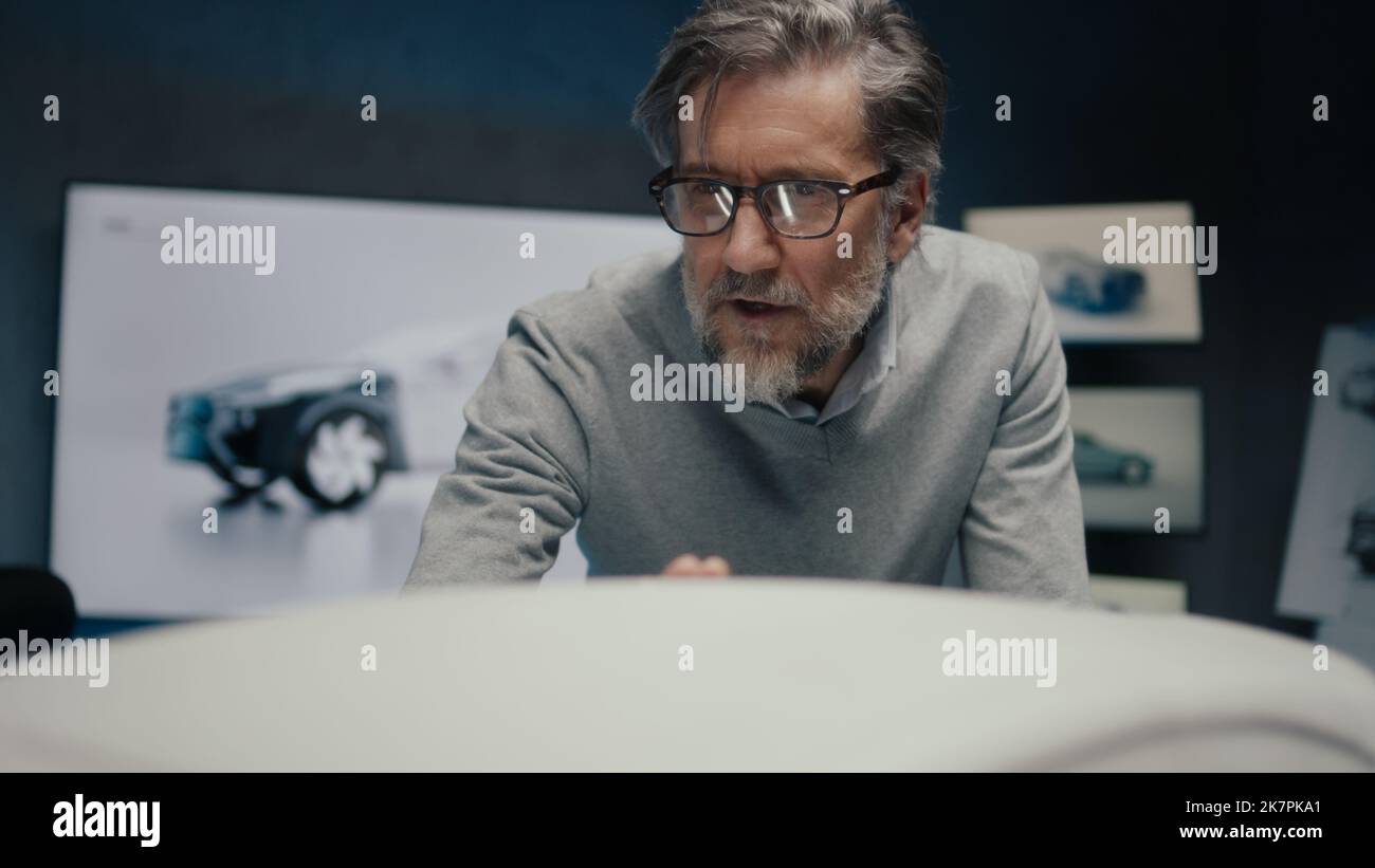Grauhaariger Automobilingenieur in einer gerahmten Brille diskutiert mit seinem Kollegen in einem Hightech-Automobilhersteller über die Designänderungen in der Skulptur eines Autos. Stockfoto