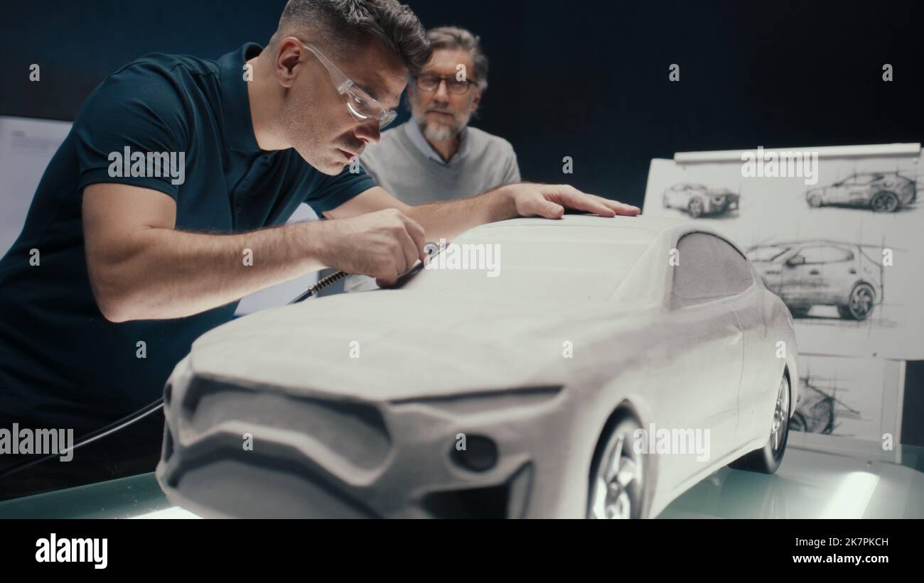 Der Automobilingenieur arbeitet mit einem rotierenden Werkzeug an der Skulptur eines Prototyps eines Automodells, während er mit seinem leitenden Kollegen diskutiert. High-Tech-Automobildesign-Unternehmen mit innovativen Technologien. Stockfoto