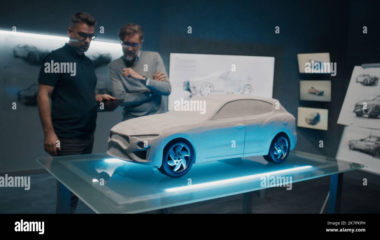 Erfahrene Automobildesigner und -Entwickler diskutieren in einem Hightech-Autorabor vor einem Rake-Modellauto-Prototyp das Design. Nehmen Sie Designkorrekturen mit einem digitalen Touchscreen-Tablet vor. Stockfoto