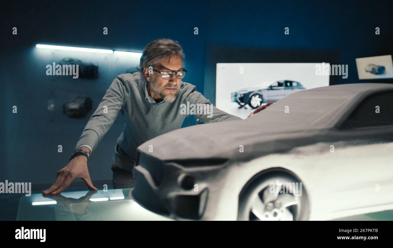 Der Automobilingenieur berührt das Modell des Prototyps aus Lehm, um endgültige Änderungen im Design vorzunehmen. Grauhaariger Autoentwickler mit gerahmten Gläsern in einem Automobilhersteller. Stockfoto