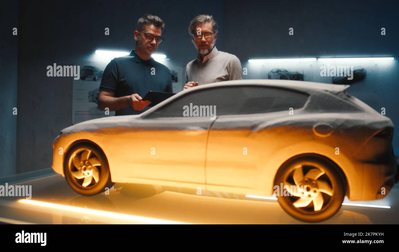 Erfahrene Automobilingenieure untersuchen das Fahrzeugmodell mit verschiedenen Lichtfarben, um endgültige Konstruktionsänderungen vorzunehmen. Verwendung eines digitalen Touchscreen-Tablets in einem Hightech-Automobilhersteller. Stockfoto
