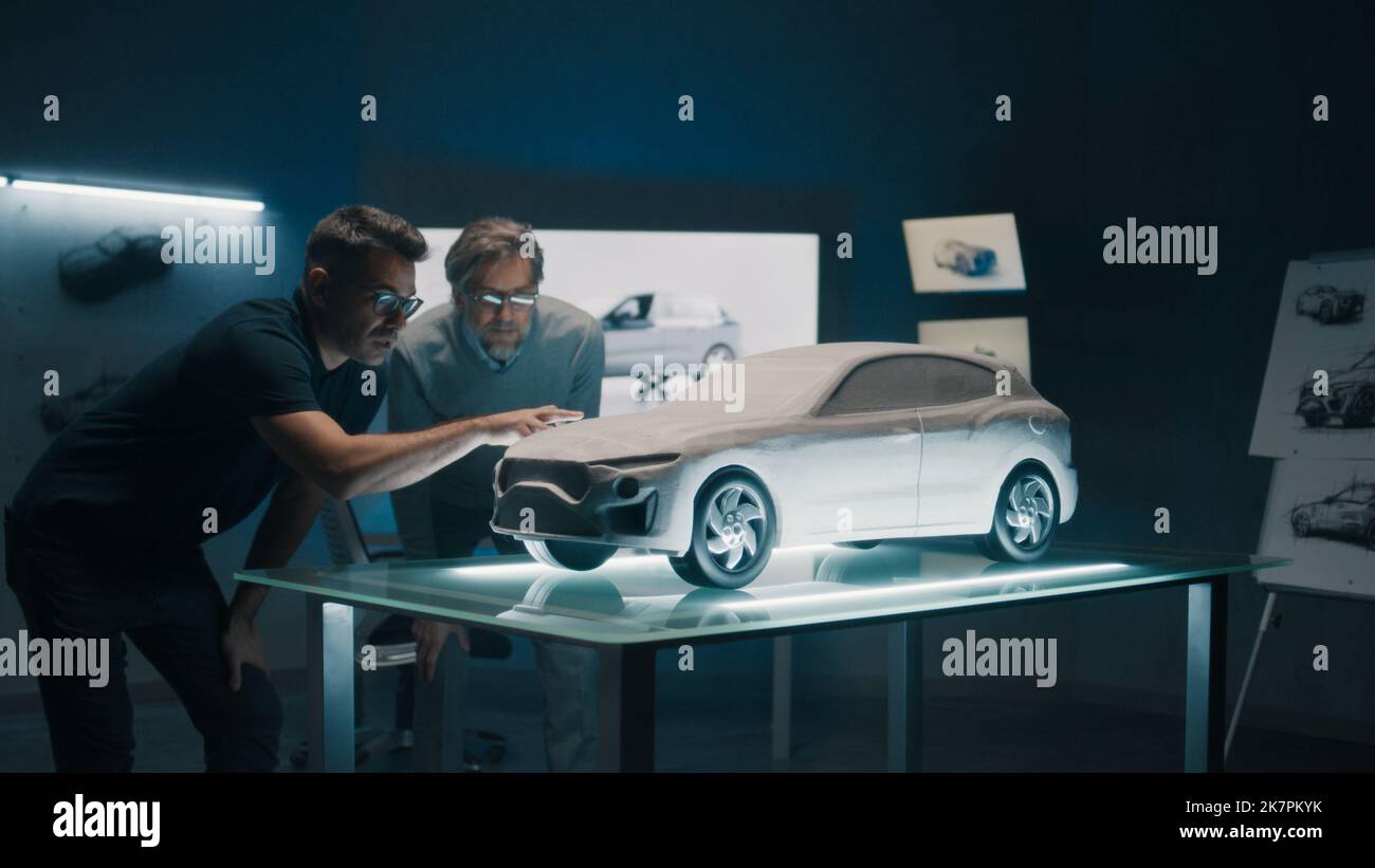 Erfahrene Automobildesigner und Autoentwickler diskutieren das Design vor einem auf einem Glastisch platzierten Rechen-Modell. Ein High-Tech-Laborraum mit LED und Skizzen. Stockfoto