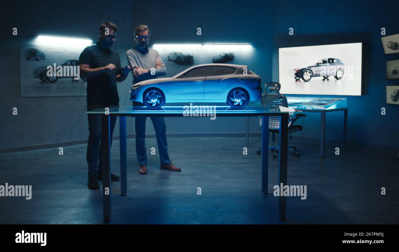 Leitende Automobilingenieure untersuchen das Prototyp-Fahrzeugmodell, das auf einem Glastisch in verschiedenen Schattierungen und Lichtfarben aufgestellt ist. Verwendung eines digitalen Touchscreen-Tablets in einem Hightech-Automobilhersteller. Stockfoto
