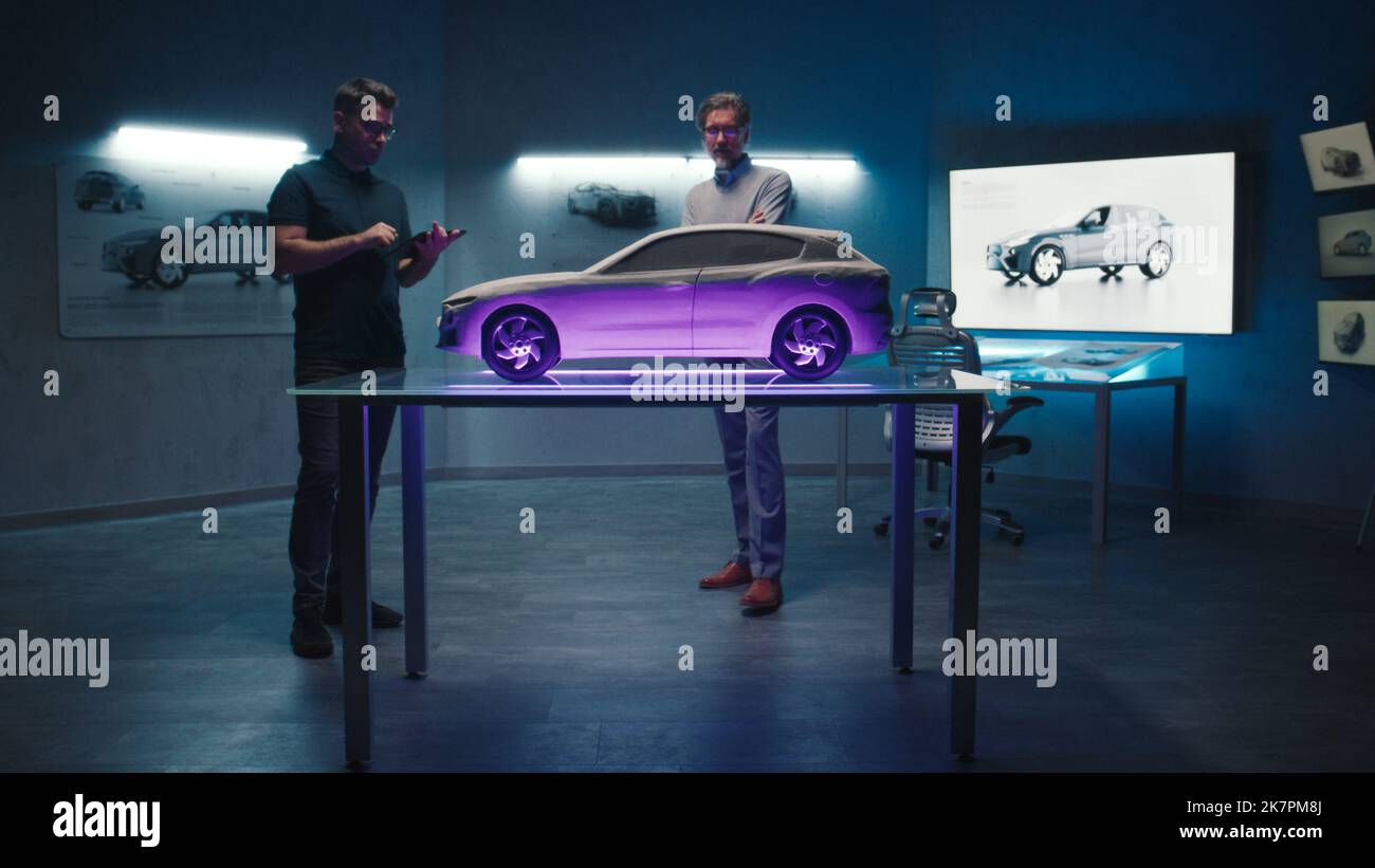 Leitende Automobilingenieure untersuchen das Prototyp-Fahrzeugmodell, das auf einem Glastisch in verschiedenen Schattierungen und Lichtfarben aufgestellt ist. Verwendung eines digitalen Touchscreen-Tablets in einem Hightech-Automobilhersteller. Stockfoto