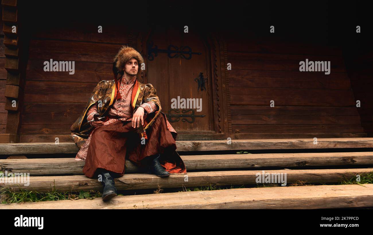 Nachdenklicher junger ukrainischer kosake in traditioneller Kleidung mit Säbel in der Hand auf der Treppe sitzend und in die Ferne schauend Stockfoto
