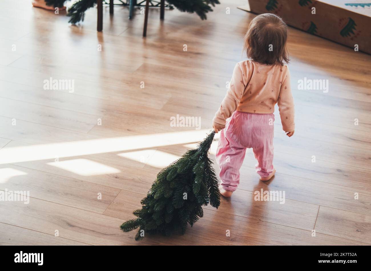 Rückansicht eines kleinen Mädchens, das einen künstlichen Christbaumzweig zu ihrer Mutter trägt, die den Baum zusammenstellt. Gemütliche festliche Atmosphäre. Warten auf Stockfoto