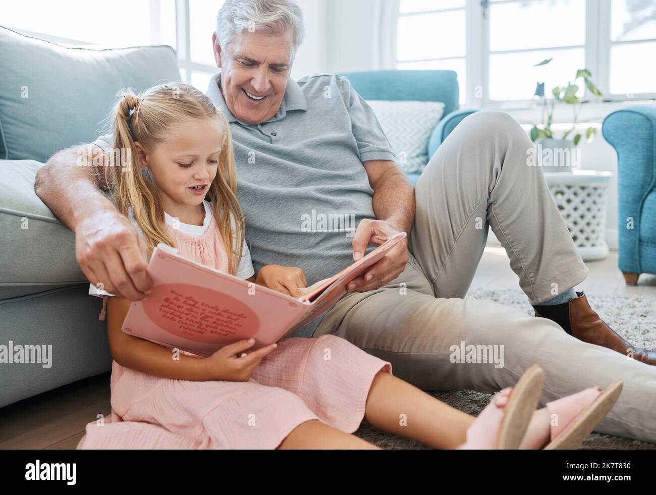 Buch, Familie und Kinder mit einem Mädchen, das ihrem Großvater auf dem Boden ihres Wohnens zu Hause vorliest. Kinder, lesen und Geschichte mit einem älteren Mann und Stockfoto