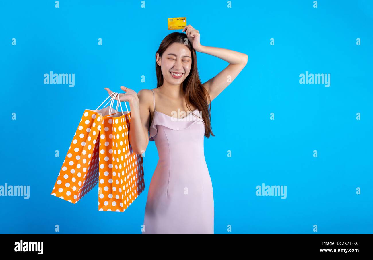 Glücklich lächelnde asiatische weibliche Shopper hält eine Reihe von Einkaufstaschen, während eine Kreditkarte hält. Modische Mädchen in Kleid Blick auf Kamera isoliert auf blauem Hintergrund - mit Kopierraum Stockfoto