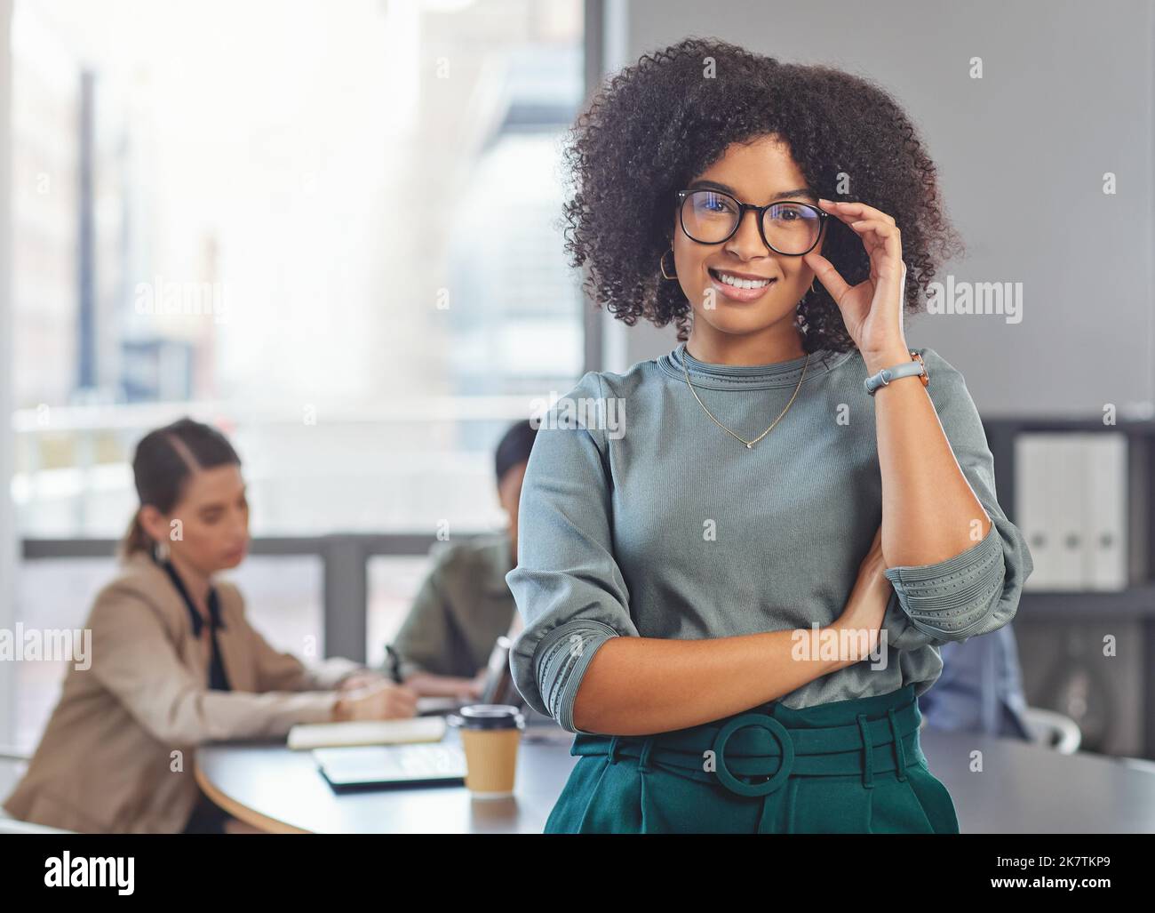 Das hält mich nicht auf. Porträt einer jungen Geschäftsfrau, die ihre Arme faltet und lächelt, während ihre Kollegen im Hintergrund ein Treffen abhalten. Stockfoto