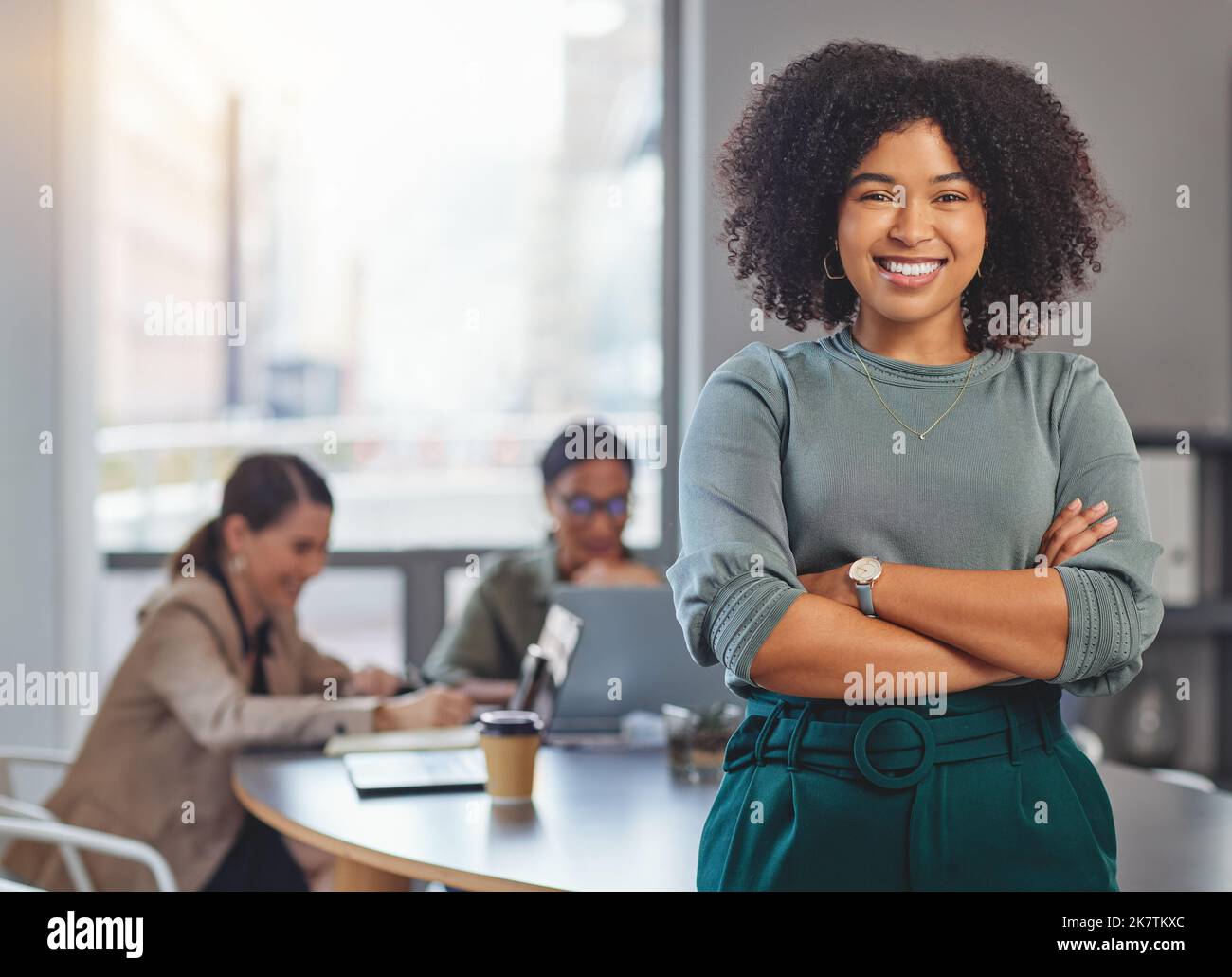 Ninja auf dein Hindernis. Porträt einer jungen Geschäftsfrau, die ihre Arme faltet und lächelt, während ihre Kollegen im Hintergrund ein Treffen abhalten. Stockfoto