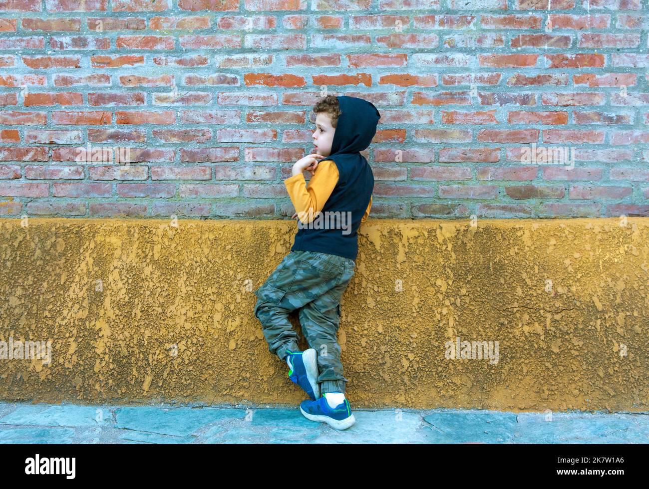 Kind in Kapuzen-Sweatshirt und Camouflage-Hosen, die an der Ziegelwand gelehnt sind Stockfoto
