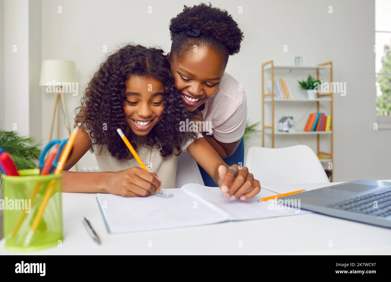 Glückliche dunkelhäutige Mutter, die ihrer fröhlichen Teenager-Tochter zu Hause beim Schreiben und Zeichnen zuschaut. Stockfoto