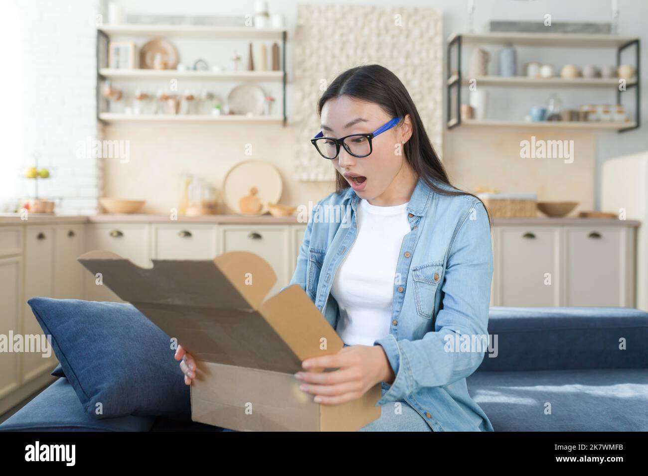 Schockiert und fröhlich Frau zu Hause erhielt einen freudigen Kauf eines Geschenks, öffnet die Box und untersucht das Geschenk mit Befriedigung, sitzt auf dem Küchensofa. Stockfoto