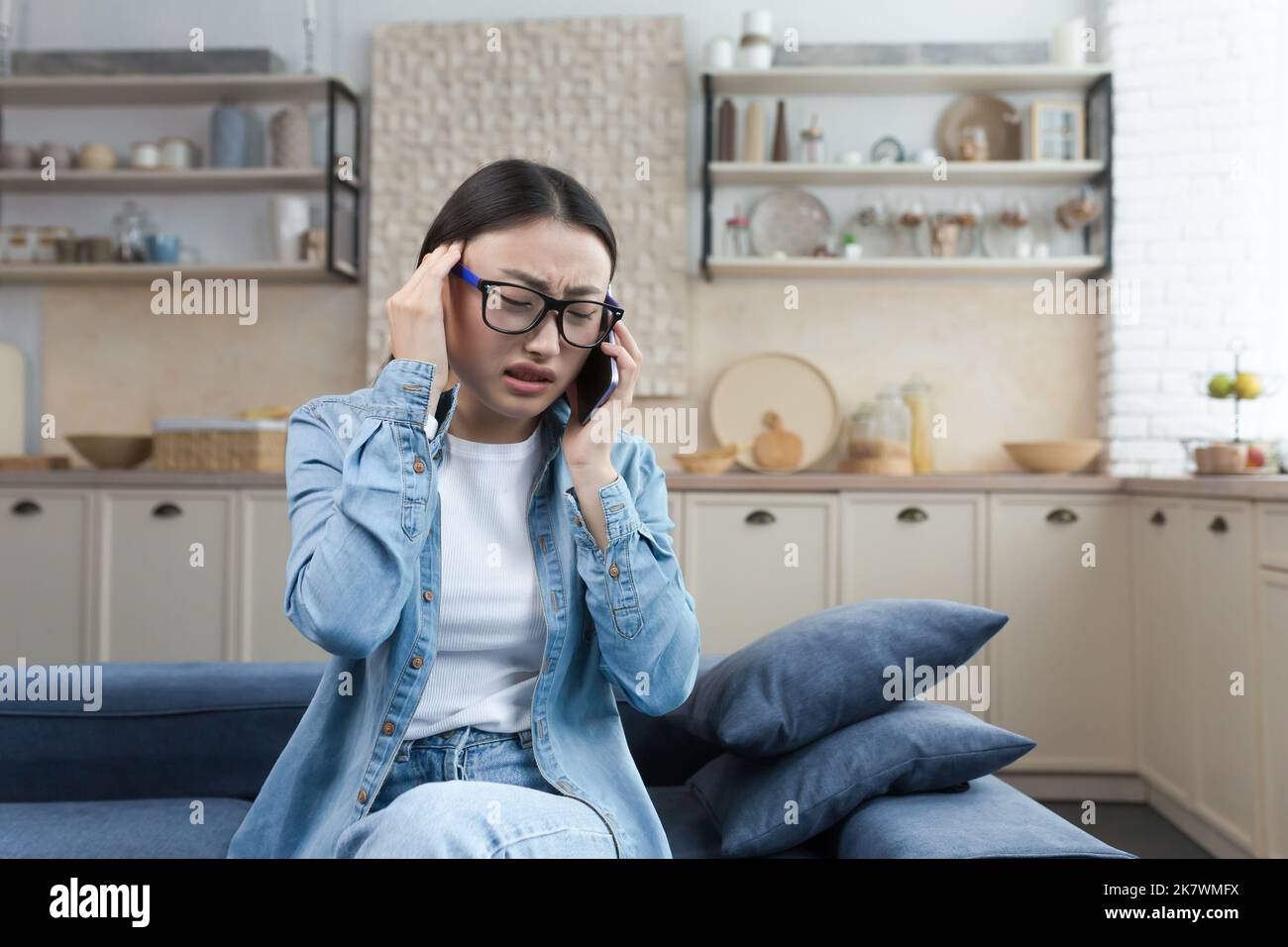 Die junge kranke Frau zu Hause hat starke Kopfschmerzen, die asiatische Frau sitzt auf dem Sofa in der Küche und spricht am Telefon, ruft den Arzt um Hilfe und konsultiert. Stockfoto