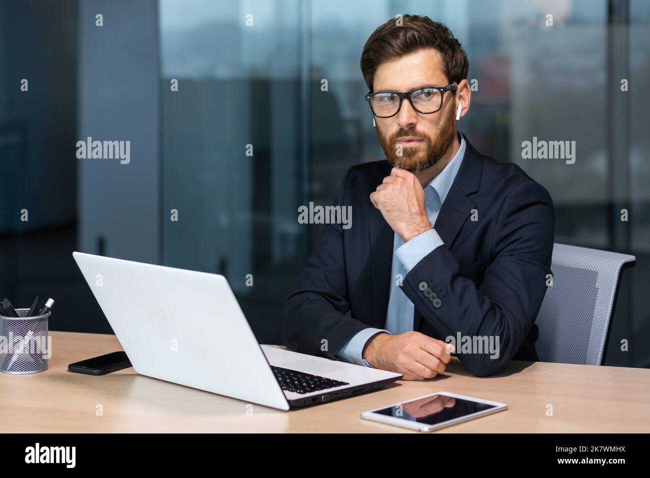 Seriös denkender reifer Geschäftsmann, der in einem modernen Bürogebäude arbeitet, Senior-Chef im Anzug und Brille, der am Laptop sitzt, Mann mit Bart, der über den zukünftigen Strategieplan nachdenkt. Stockfoto