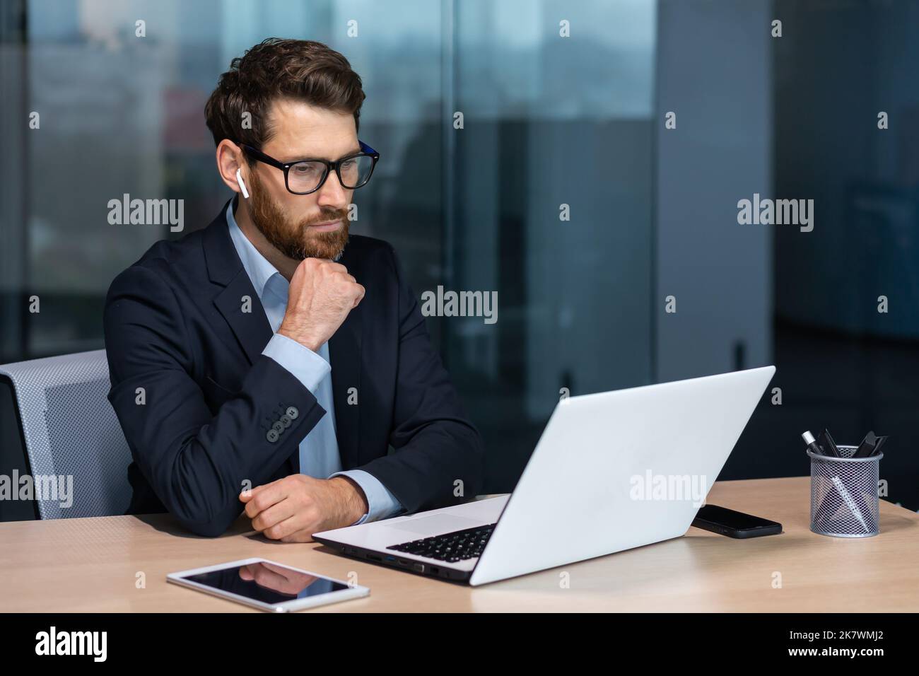 Seriös denkender reifer Geschäftsmann, der in einem modernen Bürogebäude arbeitet, Senior-Chef im Anzug und Brille, der am Laptop sitzt, Mann mit Bart, der über den zukünftigen Strategieplan nachdenkt. Stockfoto