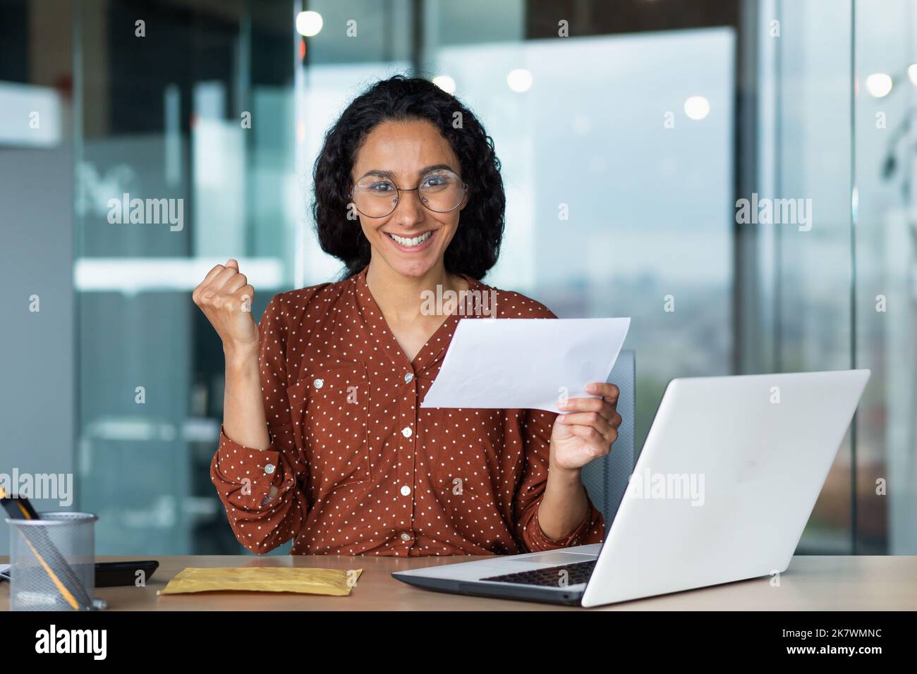 Porträt einer glücklichen Frau, hispanische Frau erhielt Brief glücklich und Blick auf die Kamera, Geschäftsfrau in Brille und lockiges Haar arbeiten im modernen Bürogebäude mit Laptop. Stockfoto