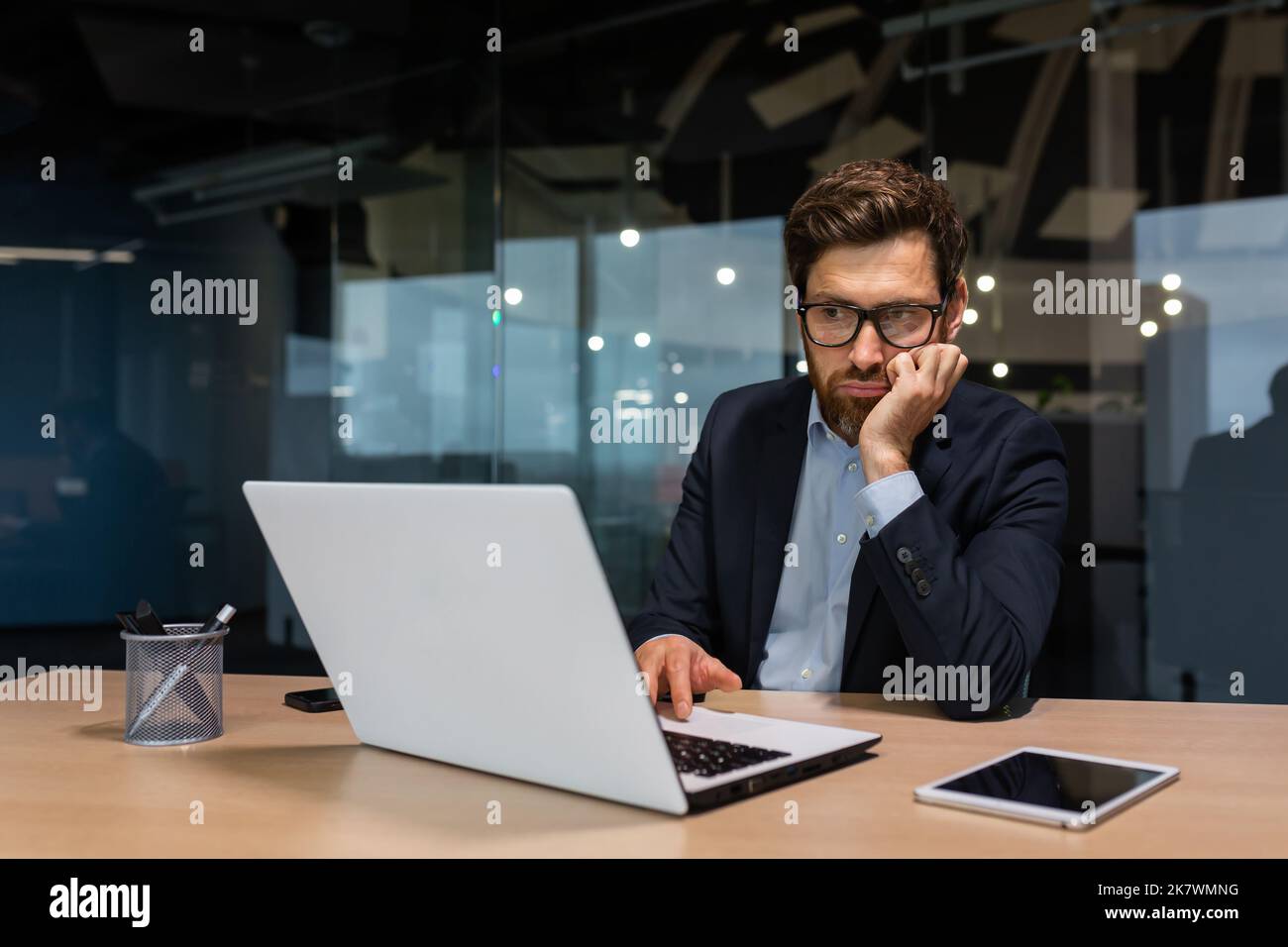 Trauriger und depressiver Geschäftsmann in Depressionen, der in einem modernen Bürogebäude arbeitet, Mann im Geschäftsanzug mit Brille und Bart, der Laptop für die Arbeit verwendet, Chef enttäuscht vom Arbeitsergebnis. Stockfoto
