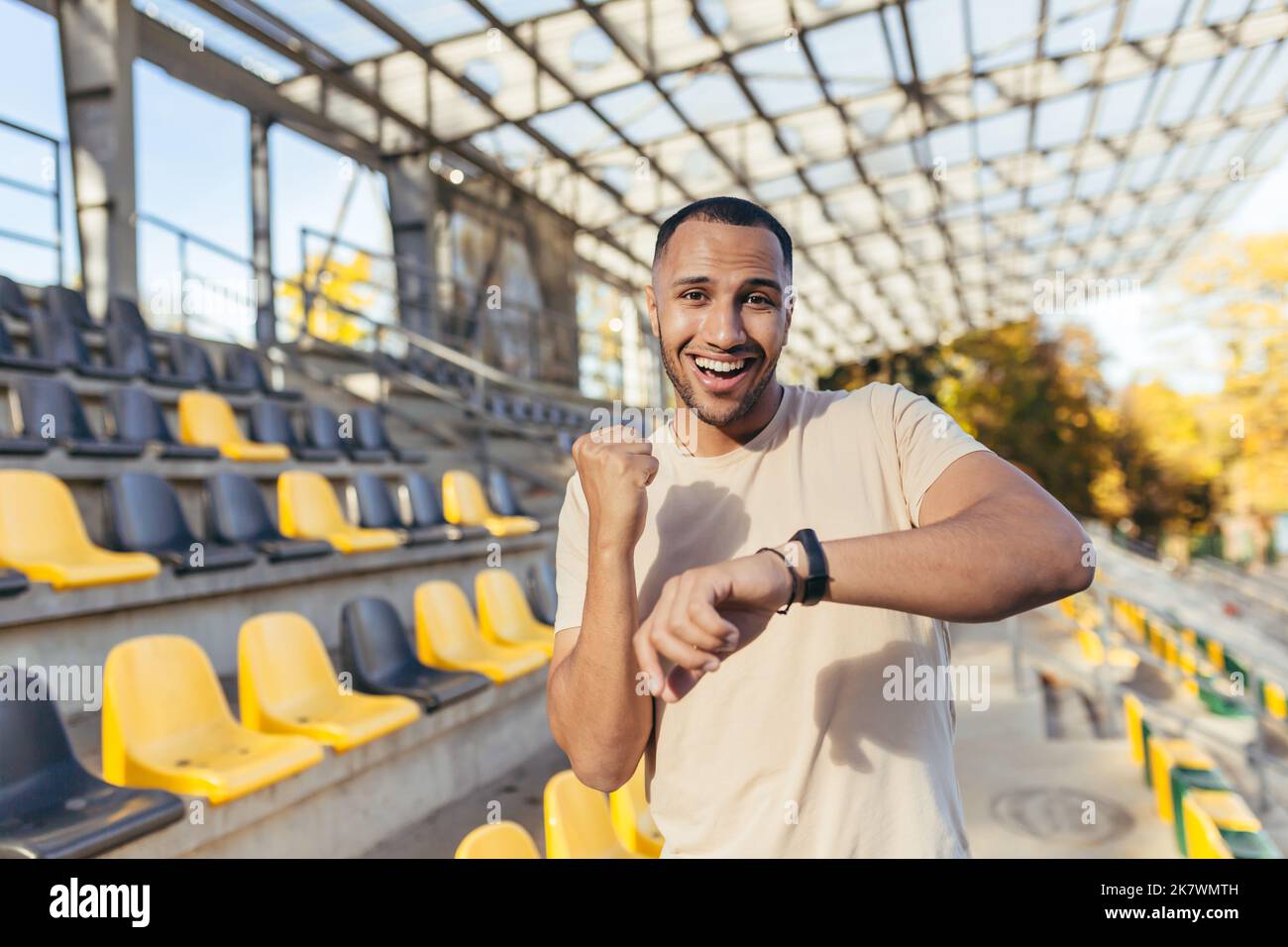 Glücklicher junger Sportler, der die Kamera anschaut und glücklich lächelt mit einem guten Fitnessergebnis, Mann, der die Zeit auf einer intelligenten Uhr des Fitnessarmbandes überprüft, tagsüber im Sportstadion läuft. Stockfoto
