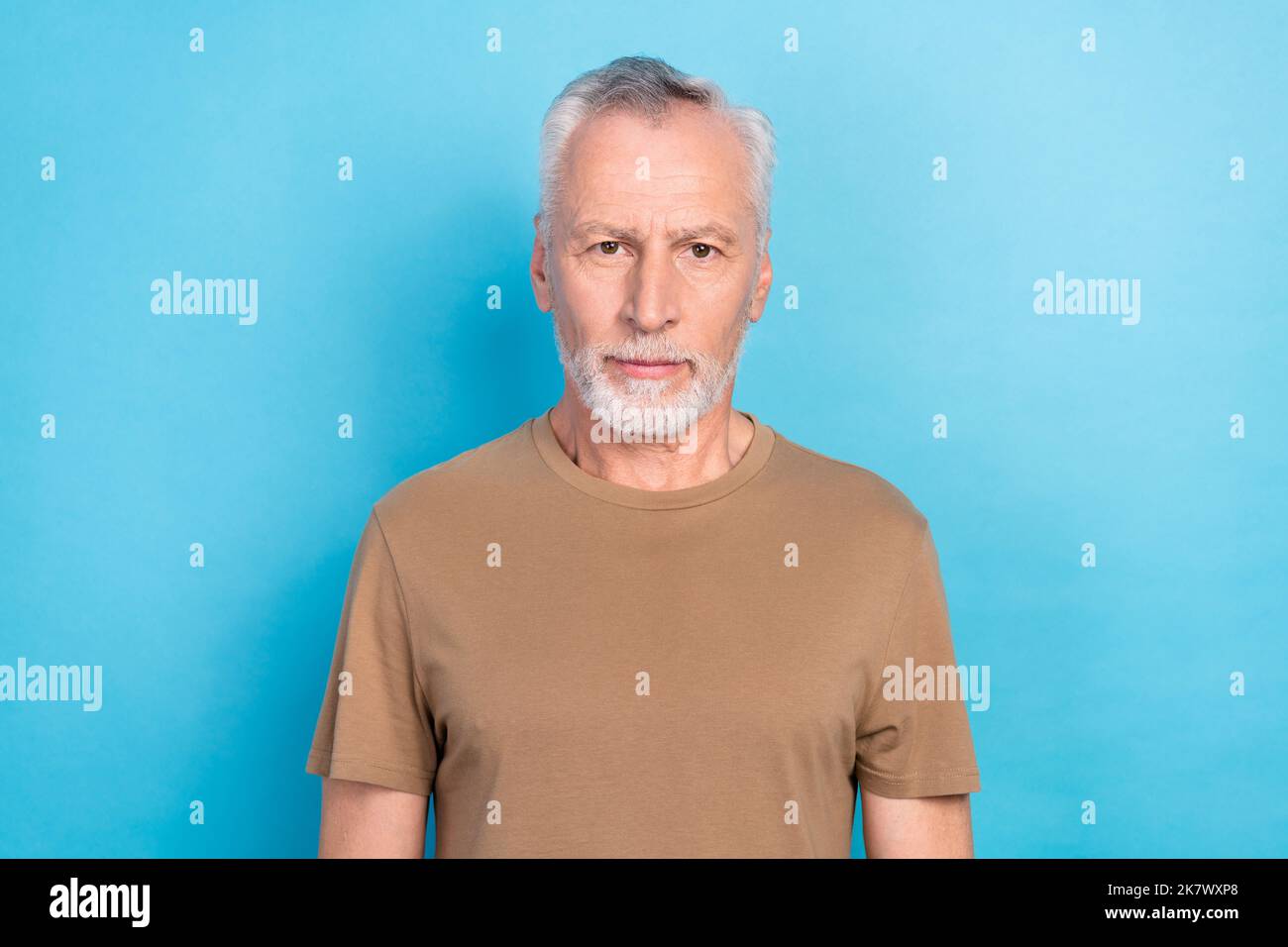 Porträt eines gutaussehenden selbstbewussten ernsten Mannes mit grauem Bart gekleidet beige T-Shirt Blick auf die Kamera isoliert auf blauem Hintergrund Stockfoto