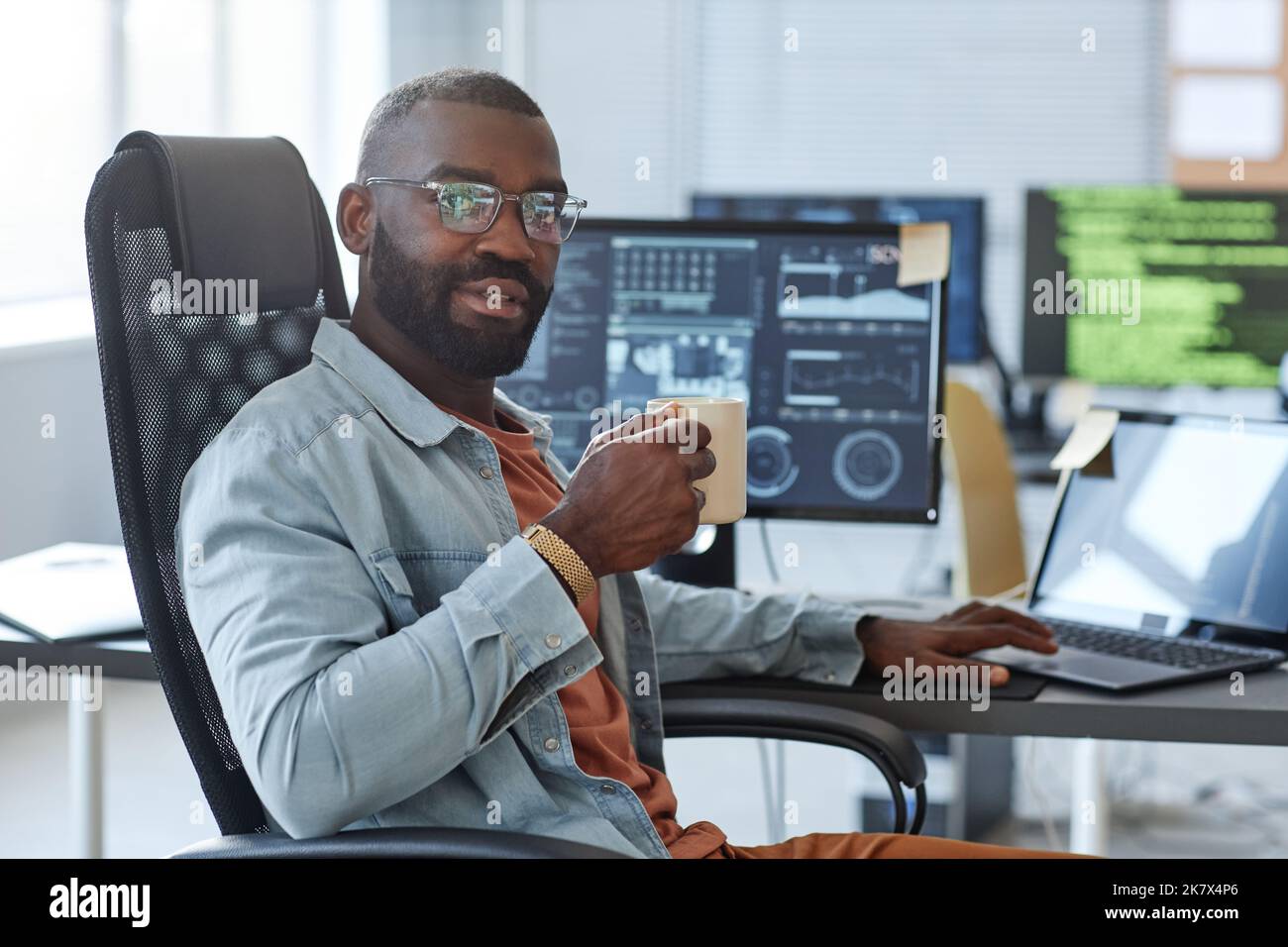 Porträt eines afroamerikanischen Mannes als Software-Ingenieur, der die Kamera anschaut, während er mit einer Kaffeetasse am Arbeitsplatz posiert und Platz kopiert Stockfoto