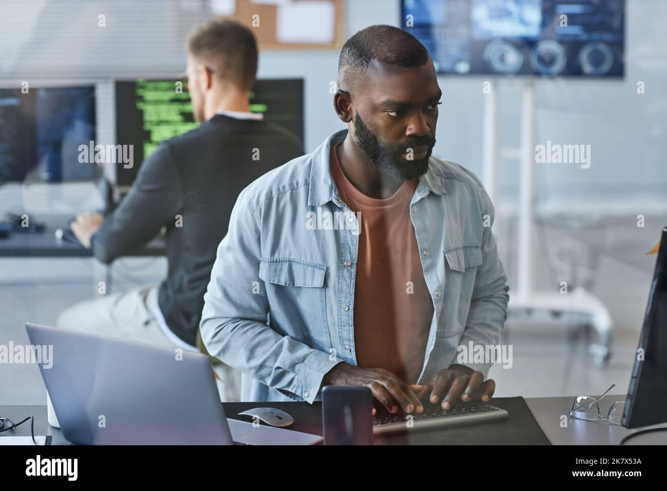 Porträt eines jungen schwarzen Mannes, der Computer verwendet, während er Software im IT-Büro eines Unternehmens hinter einer Glaswand programmiert Stockfoto