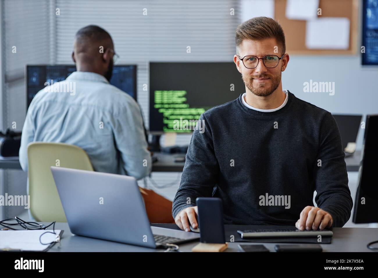 Porträt eines männlichen Softwareentwicklers, der die Kamera anlächelt, während er am Arbeitsplatz im Büro sitzt und Platz kopiert Stockfoto