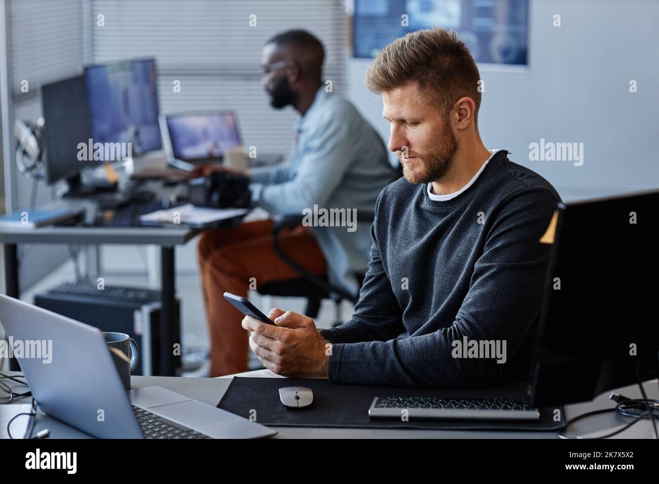Seitenansicht Porträt eines jungen kaukasischen Mannes, der am Arbeitsplatz Smartphone benutzt, während er in der Entwicklung mobiler Software arbeitet, Kopierraum Stockfoto