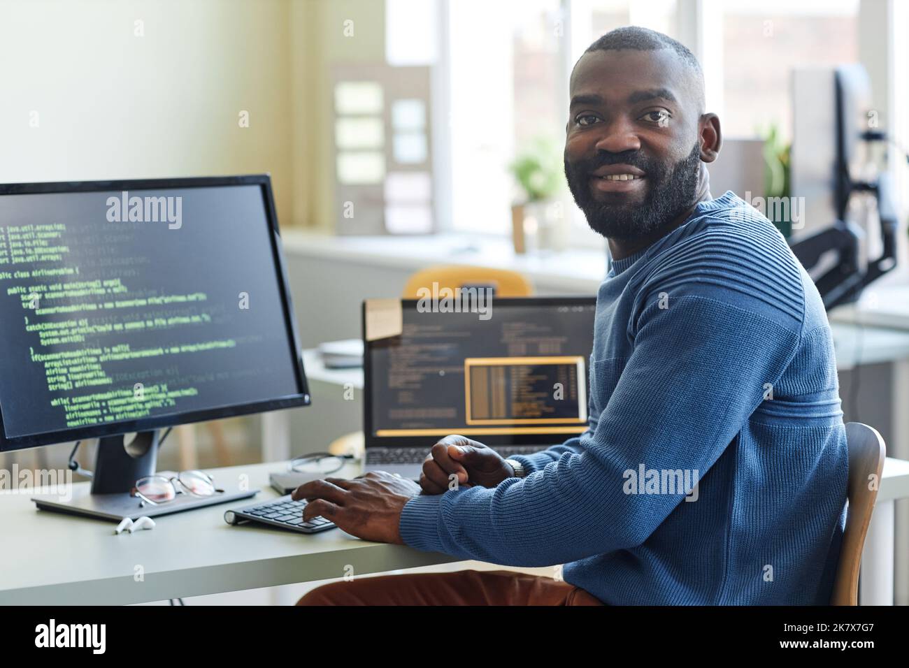 Porträt eines schwarzen Mannes als Computerprogrammierer, der beim Schreiben von Code am Büroarbeitsplatz auf die Kamera schaut Stockfoto