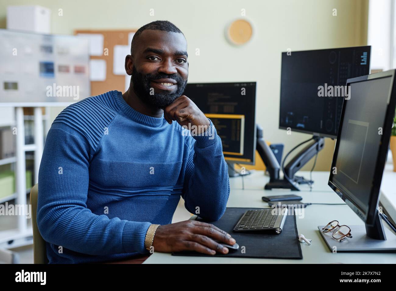 Porträt eines afroamerikanischen Mannes als Computerprogrammierer, der die Kamera anlächelt, während er am Büroarbeitsplatz Code schreibt Stockfoto