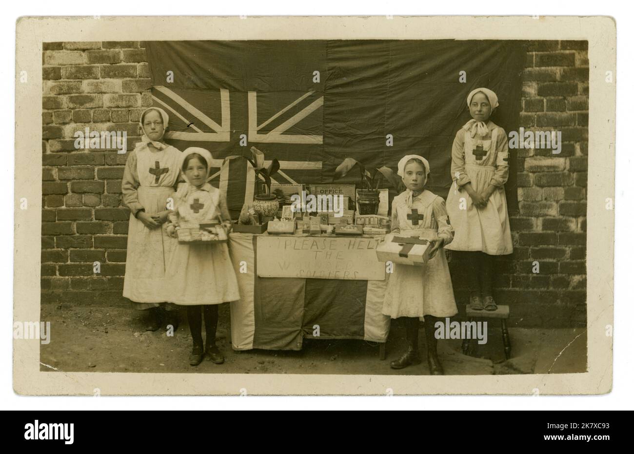 Originelle und charmante Postkarte aus der Zeit WW1 mit Kindern der Arbeiterklasse in roten Kreuzuniformen, mit einem Verkaufsstand, der ihre Spielsachen und Spiele, Süßigkeiten und Topfpflanzen verlost oder verkauft, um Geld für verwundete Soldaten zu sammeln, Union Jack-Flagge im Hintergrund, 1914-1918 Großbritannien Stockfoto