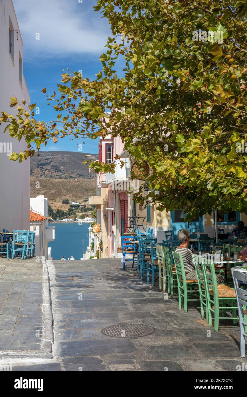 Griechisches Tavernencafe im Freien auf Chora Stadt Andros Insel mit Blick auf blaues Meer Ziel Griechenland. Die Menschen genießen die Sonne auf einer gepflasterten Gasse. Vertikal Stockfoto