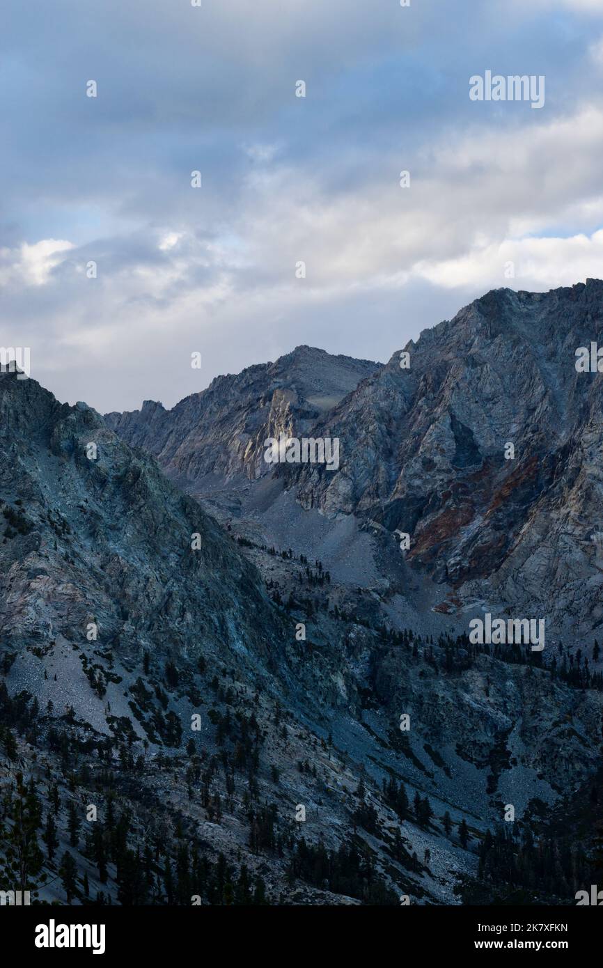 Vertikaler Blick auf dramatische Bergklippen, die in ein Tal mit verstreuten immergrünen Bäumen herabsteigen Stockfoto