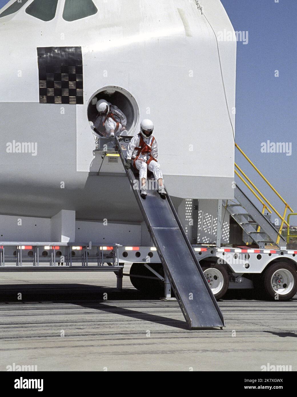 Ein freiwilliger „Astronaut“ startet während einer Rettungs- und Genesungsübung eine Ausstiegsrutsche von einem Space Shuttle-Crew-Kompartiment herunter. Während Feuerwehrleute Evakuierungsmüllchen vorbereiten, bereiten sich zwei „Astronauten“ vor, während sie während einer Rettungsübung eine Ausstiegsrutsche aus einem Shuttle-Mockup verwenden. Stockfoto