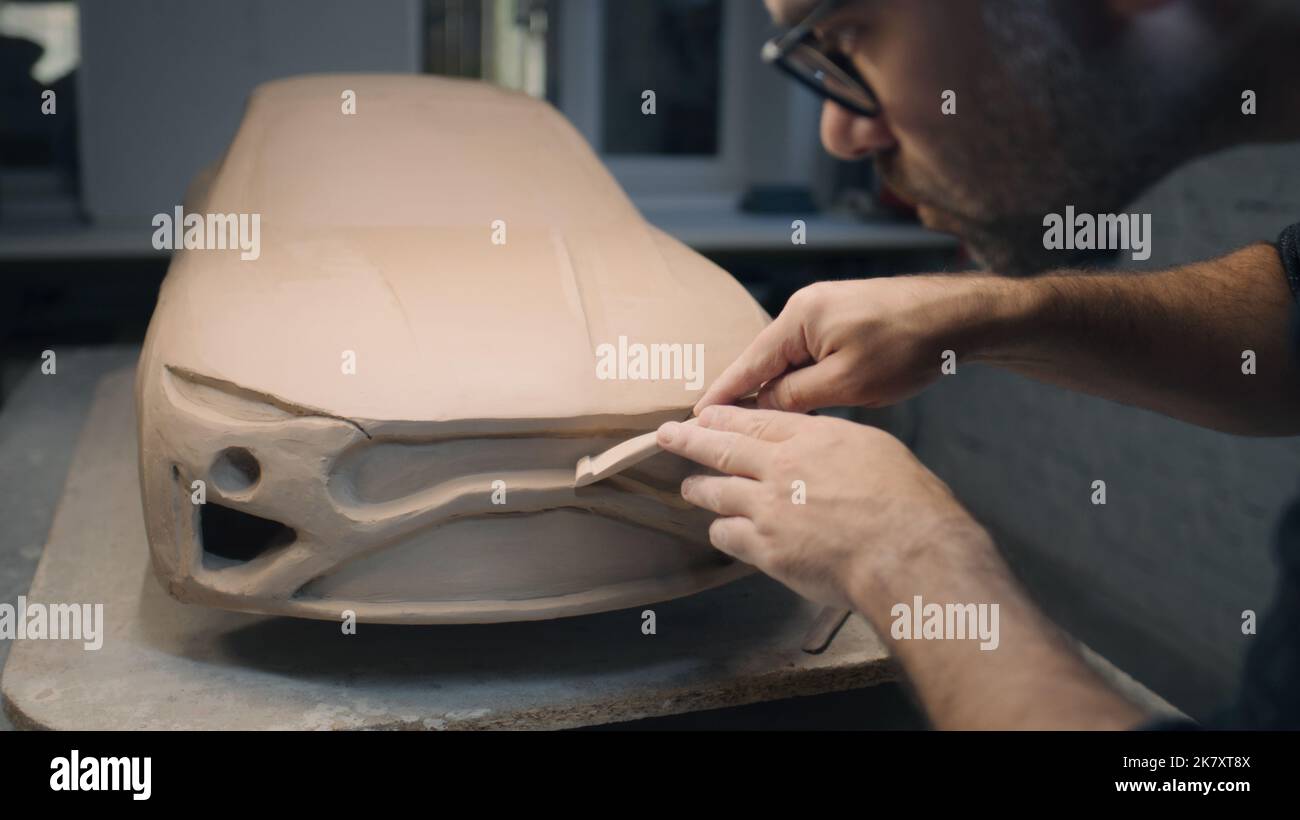 Die Hände eines männlichen Designers arbeiten an der Skulptur eines Autos mit Werkzeugen zur Bildhauerei. Glättet die Oberfläche des unfertigen Modells in einer Werkstatt. Automobilindustrie. Stockfoto