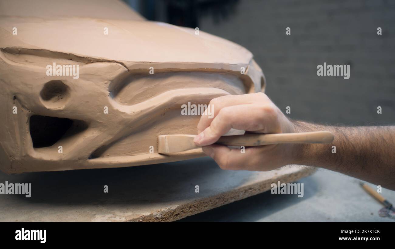 Die Hände eines männlichen Designers arbeiten an der Skulptur eines Autos mit Werkzeugen zur Bildhauerei. Glättet die Oberfläche des unfertigen Modells in einer Werkstatt. Automobilindustrie. Stockfoto