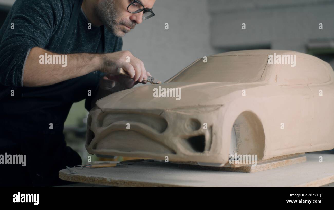 Der erfahrene Autodesigner arbeitet am Prototypenmodell eines Autos mit Holzformwerkzeugen, um die Motorhaubenoberfläche zu glätten. Unvollendetes Automodell auf einem Holztisch platziert. Stockfoto