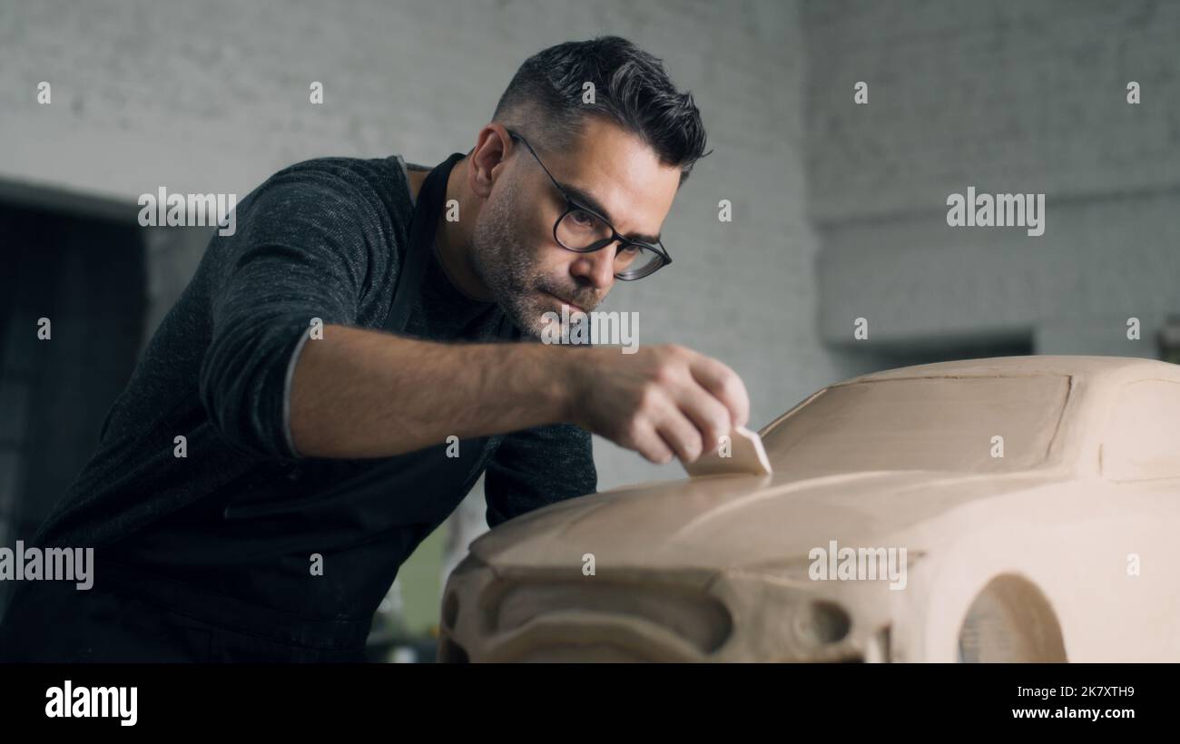 Der erfahrene Automobildesigner mit Schürze arbeitet an der Lehmskulptur des umweltfreundlichen Autos, indem er die Designdetails mithilfe eines Holzmodellierwerkzeugs schnitzt. Handgemachte Auto Skulptur aus Ton Stockfoto