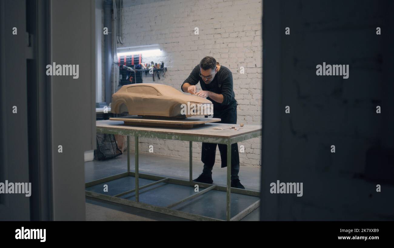 Männlicher Designer mit einer Schürze modellierenden Prototyp Modell eines Autos aus Lehm auf einem Holztisch in einer Werkstatt platziert. Verwendet Modellierwerkzeuge, um Details zu entwerfen. Stockfoto
