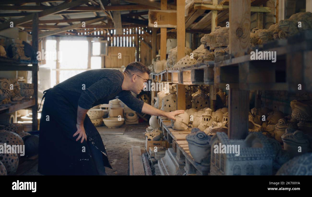 Ein Keramiker mit Schürze und schwarzen Gläsern platziert seine Kunstwerke in einem Tonstudio. Keramikwerkstatt, um seine Kunstwerke zu präsentieren. Schöne handgemachte Tonskulpturen. Stockfoto