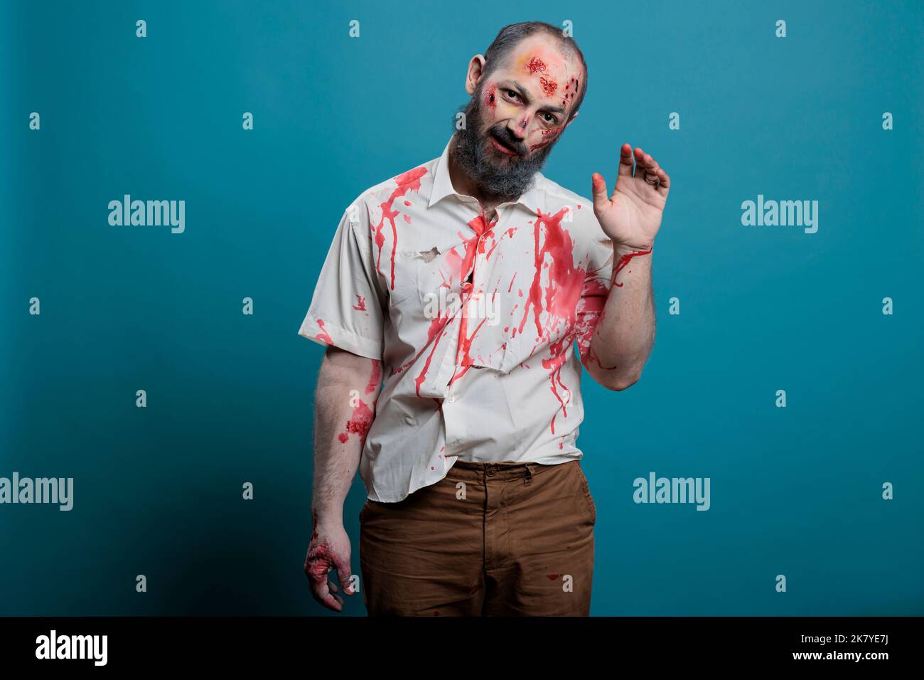 Gefährlicher Zombie winkt Hallo an die Kamera, tut Gruß Geste über blauen Hintergrund. Gruseliges, apokalyptisches böses Monster mit blutigen Narben und Wunden, das Gehirn frisst aggressive Leiche. Stockfoto