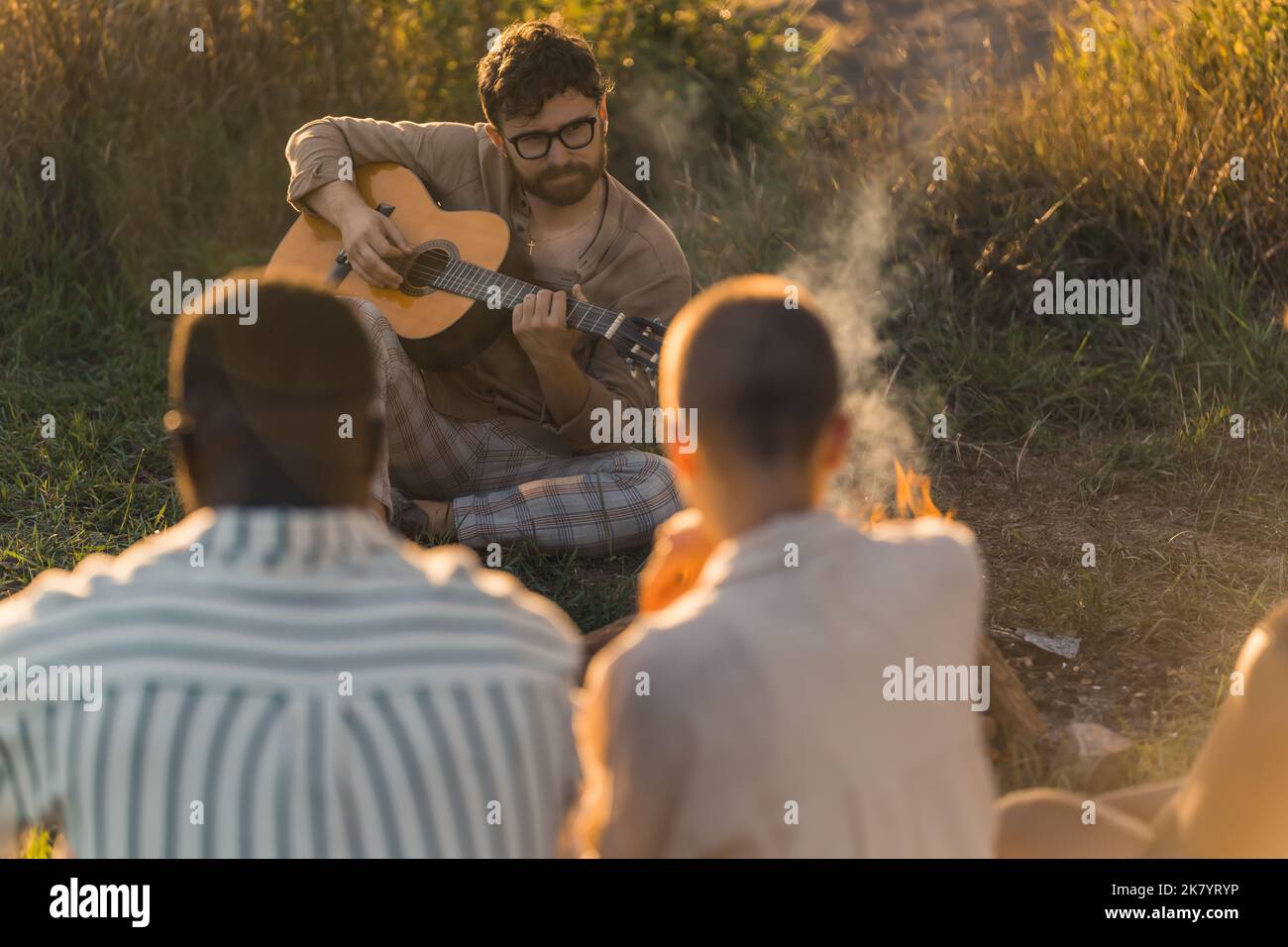 Kaukasischer Bärte kaufen in einer Brille, die vor seinen Freunden auf dem Gras sitzt und akustische Gitarre spielt. Außenaufnahme. Verschwommene Personen im Vordergrund. Hochwertige Fotos Stockfoto
