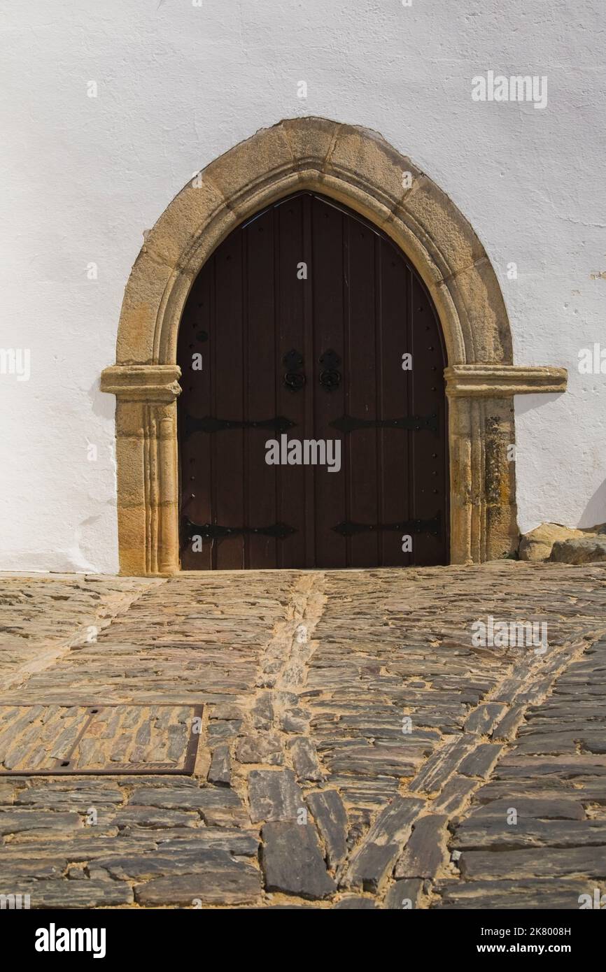 Weiß getünchtes Gebäude mit Eingangstür im gotischen Stil in einer Gasse, die mit Kopfsteinpflaster innerhalb der Mauern des Alten Schlosses in Monsaraz, Portugal, gepflastert ist. Stockfoto