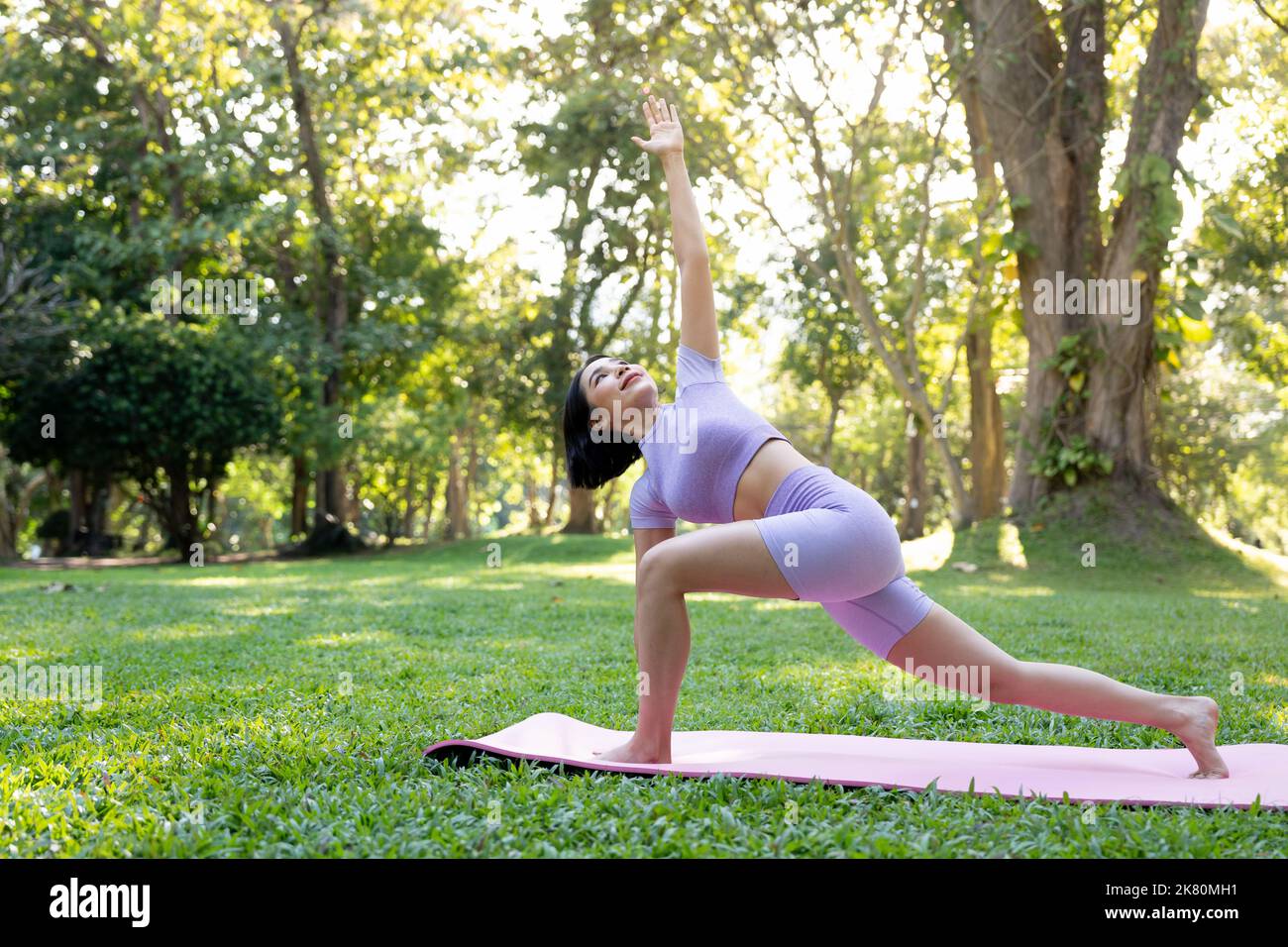 Attraktive junge asiatische Frau praktiziert Yoga, trainiert im Park, steht ein Bein auf einer Yogamatte und zeigt eine ausgeglichene Haltung. Wellness-Lifestyle und Stockfoto