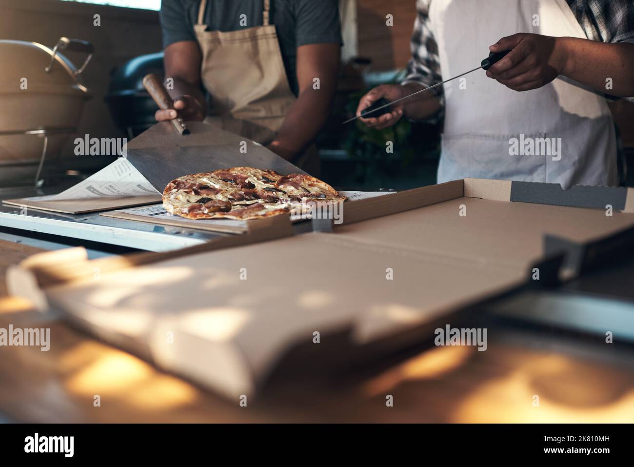 Eine heiße Pizza kommt gleich nach oben. Zwei unkenntliche Männer stehen und bereiten in ihrem Restaurant eine frisch zubereitete Pizza zum Mitnehmen zu. Stockfoto
